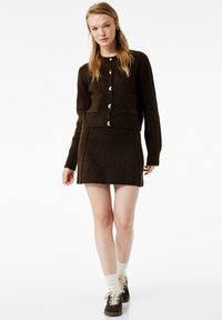 Brun strikket cardigan med rund halsudskæring og guldknapper, parret med et matchende brunt miniskørt. Modellen har hvide sokker og mørke sko på.