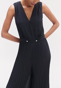 Zwarte jumpsuit met een diepe V-hals, mouwloos ontwerp, geplooide wijde benen en een tailleband versierd met twee decoratieve knopen. Gladde textuur.