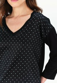 Schwarzes V-Ausschnitt-Oberteil mit hellblauen Punkten, das über eine glatte Textur und Dreiviertelärmel verfügt. Beinhaltet einen schwarzen Saum entlang des Ausschnitts.