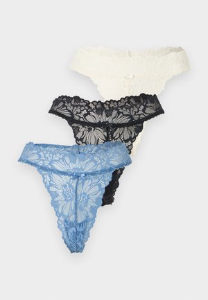 Žena nosi biely halter crop top a čierne čipkované tangá, stojí pred jednoduchým svetlosivým pozadím.