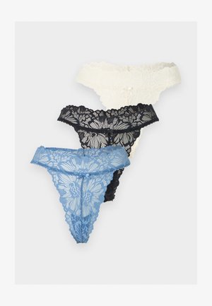 Nő fehér halter crop topot és fekete csipkés tanga alsóneműt viselve, egy sima világosszürke háttér előtt áll.