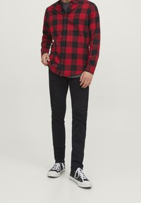 Man som bär en röd och svart rutig skjorta, mörka byxor och svartvita sneakers, stående mot en neutral bakgrund.