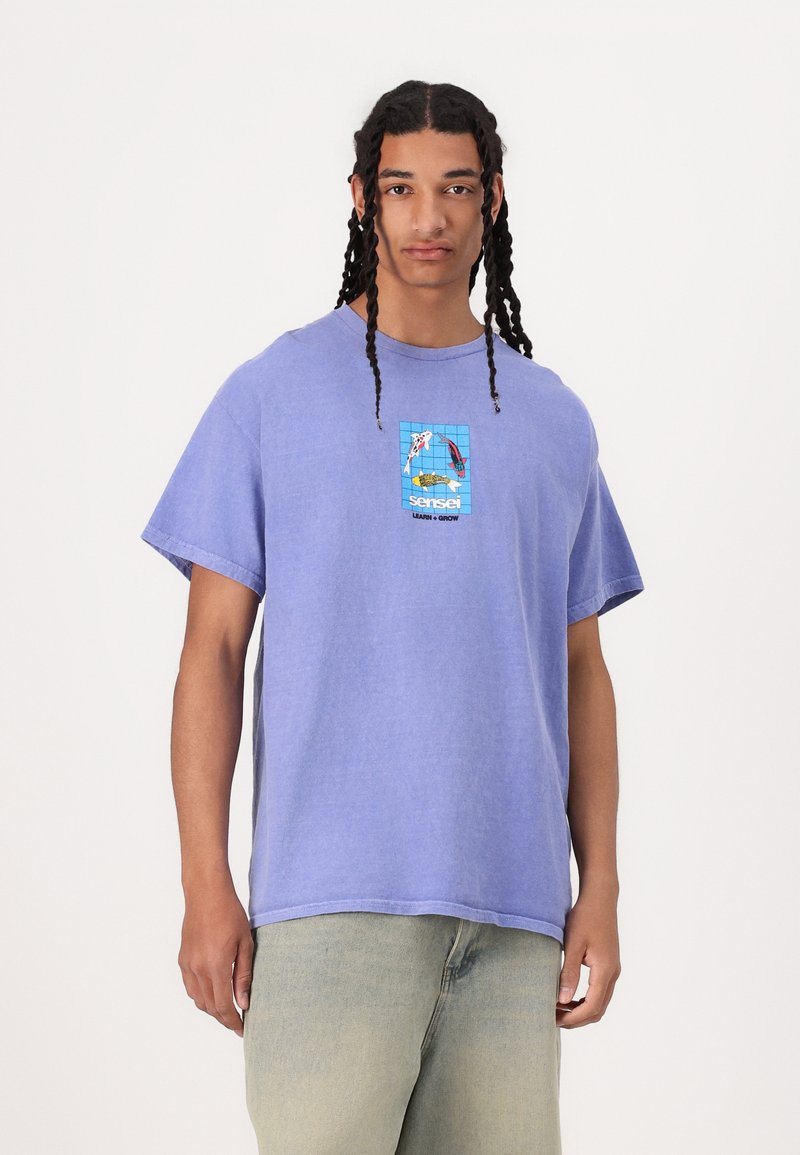 Camiseta de algodón morada de corte holgado, con cuello redondo y mangas cortas. Presenta un colorido estampado gráfico en la parte frontal, que representa un pez y una patineta.