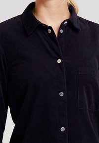 Donna che indossa una camicia a bottoni in corduroy blu scuro con bottoni a pressione argentati e una tasca sul petto, colletto visibile, maniche lunghe.