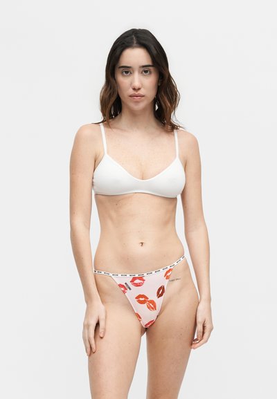 Femme portant un soutien-gorge blanc et un bas de bikini blanc avec une impression de lèvres rouges et un texte noir sur la ceinture, debout devant un fond clair uni.
