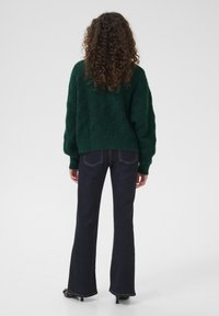 Zaļš adīts džemperis ar teksturētu rakstu, apvienots ar tumši ziliem platām biksēm un melnām apaviem ar papēdi, redzams no aizmugures.