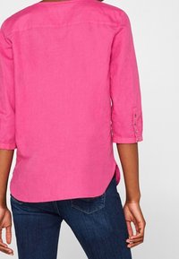Camicia lunga a maniche corte rosa in lino con maniche a tre quarti, fessure laterali e dettagli con bottoni ai polsini, indossata sopra jeans blu.