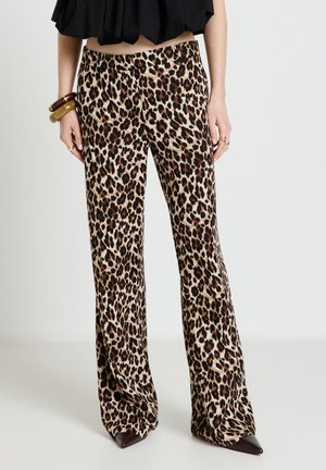 Frau mit Leoparden-Print Weitbein-Hose, einem schwarzen bauchfreien Top, spitzen dunklen Schuhen und groben Armbändern am linken Handgelenk.