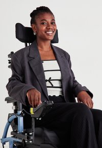 Χρήστης αναπηρικής καρέκλας με γκρίζο σακάκι πάνω από λευκό μπλουζάκι με μαύρες ρίγες. Καθισμένος σε σύγχρονη αναπηρική καρέκλα με υποστήριξη χεριών και προσκέφαλο.