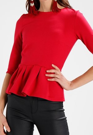 Mujer vistiendo una blusa peplum roja ajustada con mangas de tres cuartos y pantalones de cuero negros, con la mano descansando en la cadera.