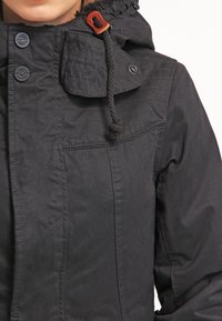 Sort hættetrøje lavet af tekstureret stof, med knaplukninger, snoretræk og quiltede detaljer på brystet.