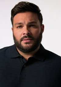 Námořnická polokošile s texturovaným pruhovaným designem, vybavená límečkem a knoflíky. Detailní záběr na muže s vousy a krátkými vlasy.