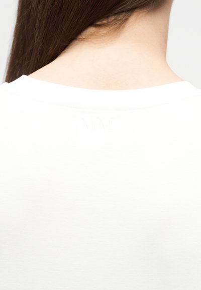 Μακριά καστανά μαλλιά που πέφτουν στην πλάτη μιας λευκής μπλούζας με στρογγυλή λαιμόκοψη, με αχνά ανάγλυφα αρχικά "MM" κοντά στη λαιμόκοψη.