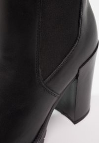 Bottine noire à talon avec une finition en cuir lisse, un panneau latéral élastique et un talon épais. Surface texturée avec des détails de couture subtils.