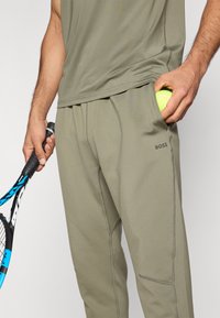 Άνετα αθλητικά παντελόνια σε ελιάς πράσινο με στενή εφαρμογή, διαθέτουν πλαϊνές τσέπες και το λογότυπο "BOSS" σε διακριτικό χρώμα. Χέρι κρατά μια μπάλα τένις και ρακέτα.
