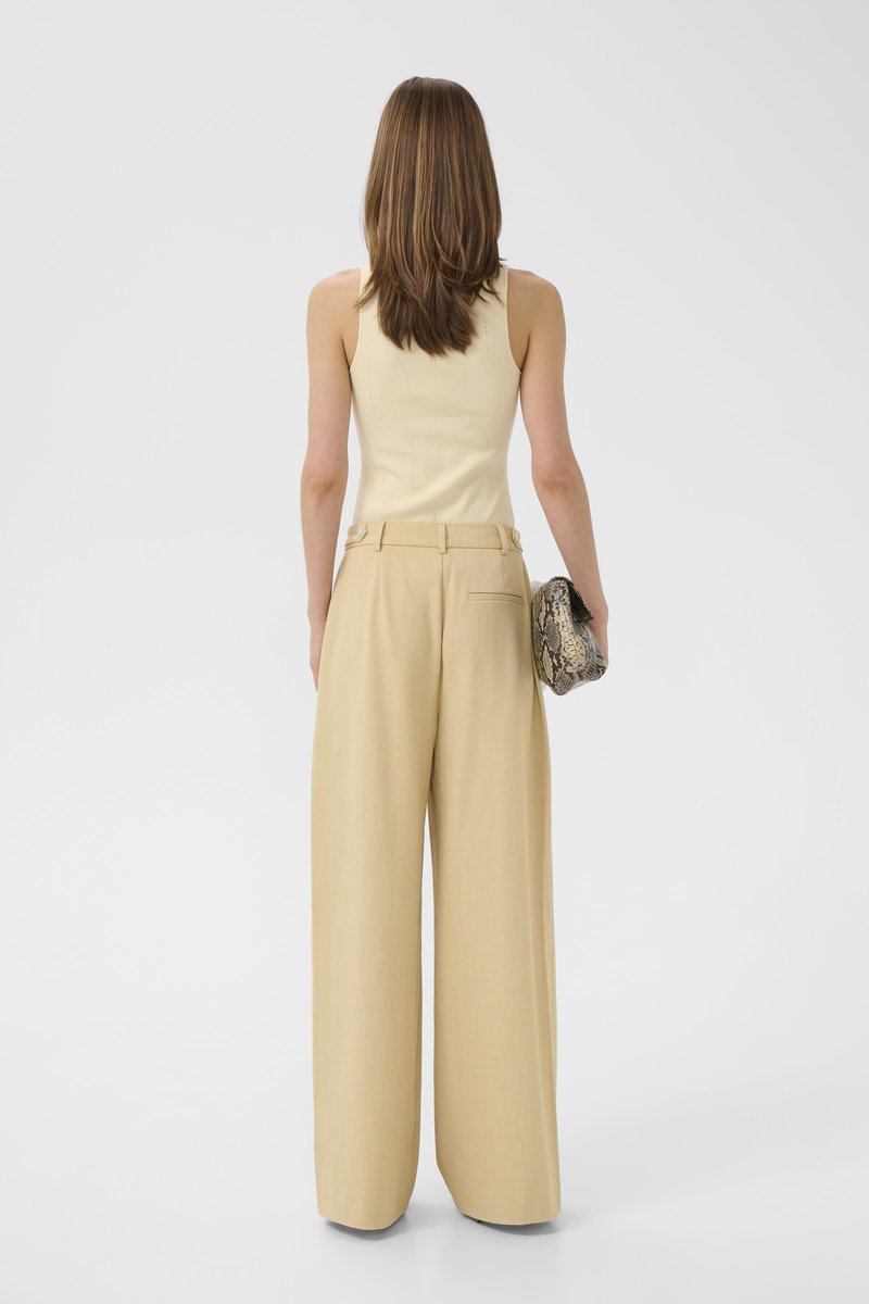 Femme debout, dos tourné, portant un haut sans manches crème, un pantalon beige à jambes larges et tenant une pochette en peau de serpent dans sa main droite.