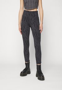 Person klädd i högmidjade mörkgrå och svarta zebramönstrade leggings med svarta snörade plattformstövlar mot en enkel vit bakgrund.