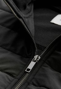 Gros plan d'une veste noire partiellement ouverte avec des dents de fermeture éclair en métal visibles et une languette de tirage.