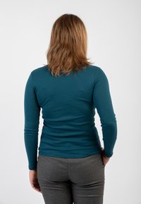 Maglietta a maniche lunghe color teal realizzata in un tessuto morbido, design aderente, abbinata a pantaloni grigi aderenti, mostrata da dietro.