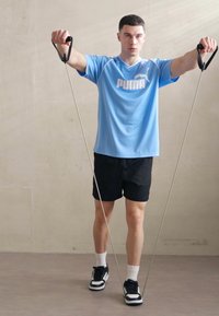 Blå trænings T-shirt med hvidt logo, sorte shorts og sorte sneakers. En person holder modstandsbånd og står på et lyst gulv.