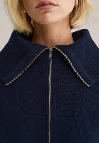 Námořnický svetr na zip s výraznou ribovanou texturou, širokým límcem a zlatým zipem, který zdůrazňuje moderní design.