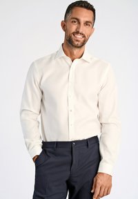 Camicia bianca a maniche lunghe con colletto classico e chiusura a bottoni, abbinata a pantaloni scuri, che presentano una superficie liscia e una vestibilità sartoriale.