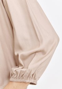 Bluzka z długim rękawem z marszczonymi mankietami, wykonana z miękkiego beżowego materiału, charakteryzująca się luźnym krojem i gładką fakturą. Brak widocznych wzorów lub akcentów.