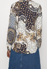 Camicia a maniche lunghe con una combinazione di motivi paisley e floreali in blu navy, bianco e oro. Tessuto leggero con dettaglio sulla schiena con bottoni.