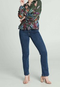 Blusa verde con motivo floreale e maniche lunghe, abbinata a jeans blu e sandali beige con tacco a blocco. Caratterizzata da colori vivaci e un tessuto morbido.