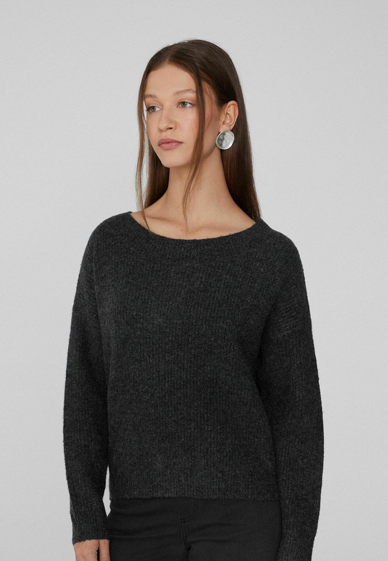 Dunkelgrauer gestrickter Pullover mit weitem Ausschnitt, langen Ärmeln und strukturierter Oberfläche. Kombiniert mit schwarzen Hosen, die ein einfaches, lässiges Design aufweisen.