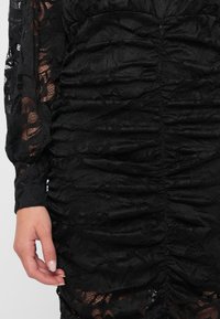 Μαύρο φόρεμα από δαντέλα με ραμμένο υφή, μακριά μανίκια και φλοράλ σχέδιο. Λείο ύφασμα με διαφανείς λεπτομέρειες και εφαρμοστή σιλουέτα.