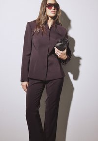 Frau trägt einen dunkel maßgeschneiderten Anzug, gro ße Sonnenbrille, hält eine schwarze Clutch-Tasche, steht vor einem weißen Hintergrund mit Schatten.