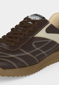 Sneakers marron et beige avec surpiqûres décoratives blanches, lacets marron et semelle en caoutchouc à motif rainuré.
