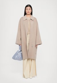 Beige oversized wool coat with a collar and zipper, paired with a cream sweater and wide-leg pants. Holding a light blue bag.