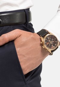 Goldfarbene Chronograph-Uhr mit einem schwarzen Lederarmband und einem dunklen Zifferblatt, ausgestattet mit drei Ziffernblättern und goldenen Akzenten, getragen an einer Hand.