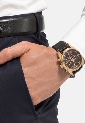 Goldfarbene Chronograph-Uhr mit einem schwarzen Lederarmband und einem dunklen Zifferblatt, ausgestattet mit drei Ziffernblättern und goldenen Akzenten, getragen an einer Hand.