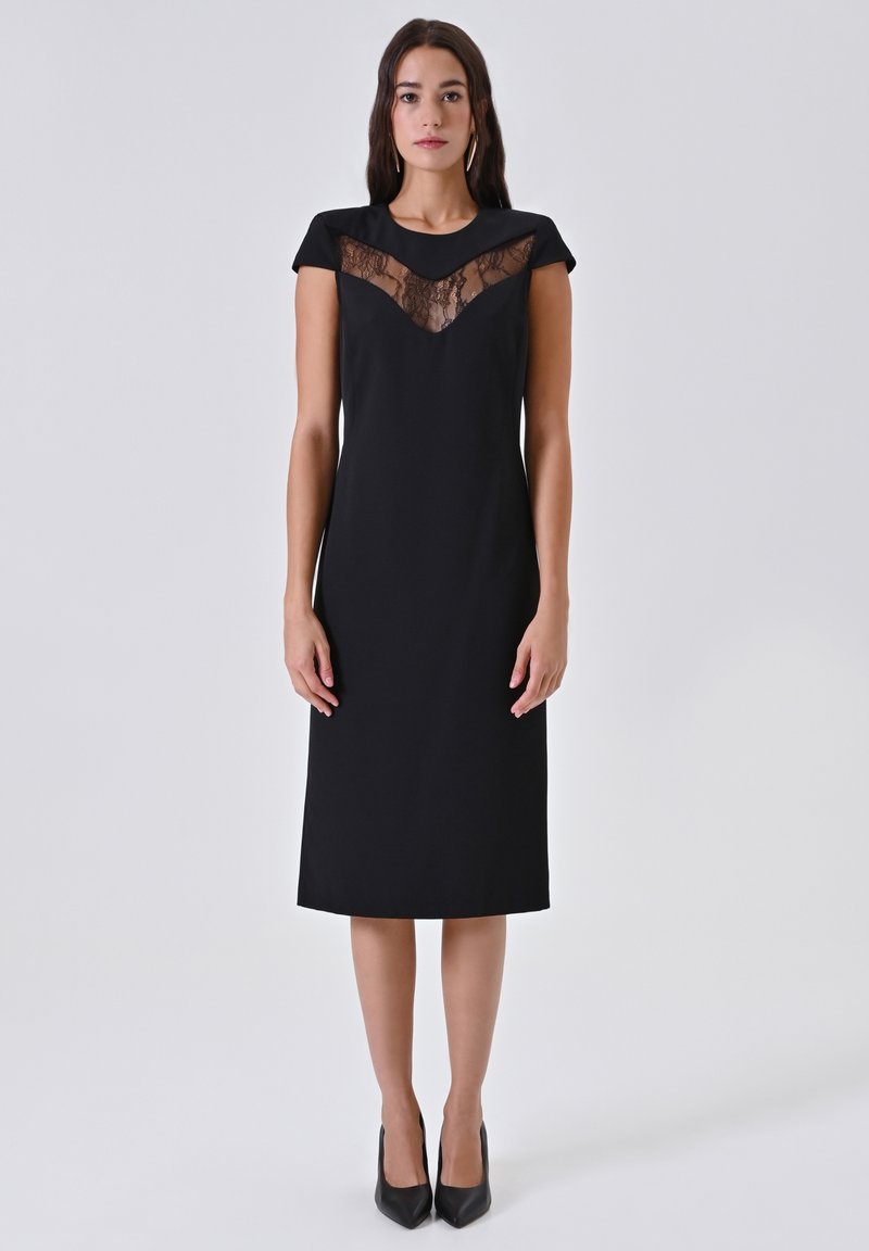 Donna che indossa un vestito nero a lunghezza ginocchio con dettagli in pizzo sul petto, in piedi con le mani rilassate e indossando tacchi a punta neri.