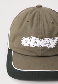 Khaki värvi pesapalli müts valge broderinguga "obey" logo, kontrastne roheline ääre ja valge voodriga servade ääres. Puuvillane kangas nähtava tekstuuriga.