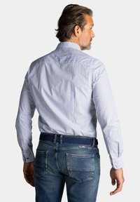 Langetermijn blauwe blouse met een wit stippenpatroon, gecombineerd met blauwe jeans. Voorzien van knoopmanchetten en een geweven riem. Zichtbare details aan de achterkant.