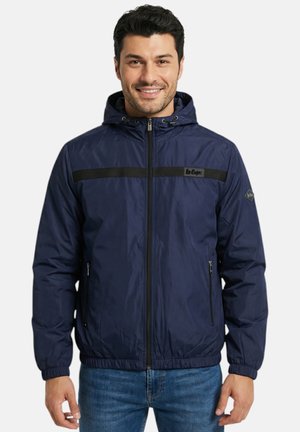 Homme souriant portant une veste à capuche zippée bleu marine avec une bande horizontale noire et des poches latérales zippées, associée à un jean bleu.