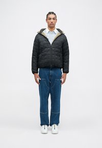 Black quilted puffer jacket with a hood and olive green lining. The model wears blue wide-leg jeans and white sneakers.