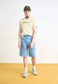 Beige T-kreklu ar 'Alpha Industries' logo, zilo džinsus šortus un pelēkzaļu sporta apavu. Aksesuāri ir zaļš beisbola cepure.
