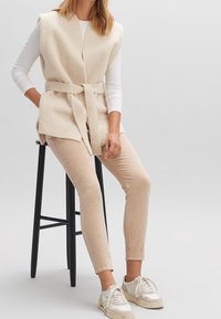 Gilet beige senza maniche, testurizzato e con una fascia, indossato sopra una maglietta bianca a maniche lunghe. Pantaloni attillati beige chiaro e sneaker beige completano l'outfit.