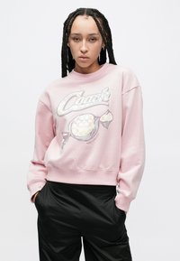 Ανοιχτό ροζ φούτερ με λαιμόκοψη crew και χαλαρούς ώμους, που διαθέτει μια λαμπερή γραφική εκτύπωση λογότυπου και σχέδιο ζαχαρωτού. Μαλακό υλικό με άνετη εφαρμογή.