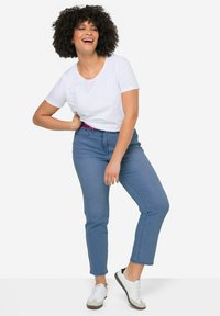 Camiseta blanca de canalé, jeans de tiro alto color azul claro y zapatos grises con cordones blancos. Notable cinturón púrpura como acento. El modelo posa con la mano en la cadera.