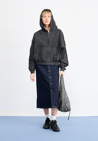 Sweat à capuche noir zippé avec motif texturé, assorti à une jupe boutonnée en denim. Accessoires comprenant un sac à motifs et des baskets noires.
