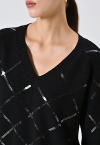 Maglione nero lavorato a maglia con scollo a V, caratterizzato da un motivo diagonale di paillettes argento disposte a griglia sulla parte anteriore.