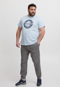 Cămiză din bumbac de culoare albastru deschis cu un imprimeu grafic bleumarin care citește "Original Blue Denim", asortată cu pantaloni casual gri și adidași bleumarin.