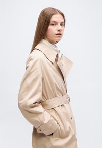 Νέα γυναίκα με ίσια μαλλιά που φοράει ένα μπεζ καμπαρντίνα με ζώνη, στέκεται προφίλ με τα χέρια στις τσέπες του παλτού, μπροστά σε μονόχρωμο φόντο.