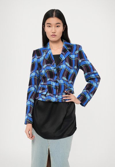 Blazer blu e nero con motivo a stampa e design su misura, dotato di colletto, bottoni e silhouette aderente. Indossato sopra una maglia nera e una gonna di jeans.